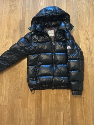 Svart dunjacka från Moncler - Snygg svart dunjacka från Moncler med glansig finish, huva och dragkedja framtill. Jackan har Moncler-logga på ärmen, praktiska fickor med dragkedja och quiltad design. Perfekt för kalla vinterdagar och riktigt trendig look.