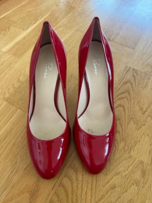Röda lackpumps från Rizzo strl 36 (som ny) - Eleganta röda lackpumps från Rizzo i storlek 36. Klassisk och tidlös modell med feminin siluett och smal klack på ca 8–9 cm.  Skorna är tillverkade i Italien med lädersula (Vero Cuoio) och har en vacker djup röd färg som passar både fest, middag, bröllop och andra speciella tillfällen.   I fint skick som ny, används fåtal gånger inomhus