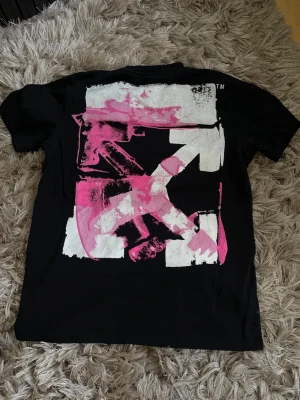 Svart Off-White t-shirt med tryck - Svart t-shirt från Off-White med ikoniskt stort grafiskt tryck i vitt och rosa på ryggen samt mindre tryck framtill. Klassisk passform och rund halsringning. Perfekt för dig som gillar streetwear och unika detaljer.