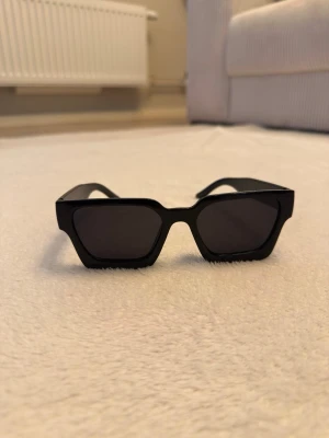 Svarta kantiga solglasögon - Snygga solglasögon med breda, svarta bågar och mörka glas. De har en modern, kantig form som ger ett coolt intryck och passar perfekt för dig som vill sticka ut. Materialet är syntet och designen är stilren och trendig. Oanvända, unisex