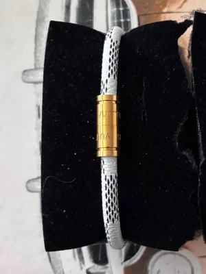 Vitt armband med guldmagnetlås - Snyggt armband i vitt med svartmönstrad textil och ett magnetlås i guldfärgad metall. Låset har diskret gravyr och armbandet har en modern, minimalistisk look som passar till många stilar. 20cm
