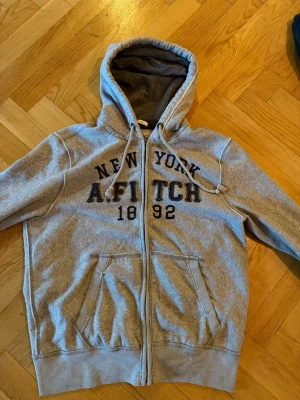 Grå hoodie från Abercrombie & Fitch - Snygg grå zip-hoodie med tryck 'New York A.FITCH 1892' på bröstet. Mjuk insida, två stora fickor fram och dragsko i huvan. Perfekt för en avslappnad streetstyle-look.