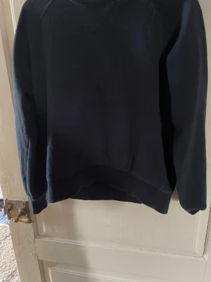 Svart enkel hoodie - En svart hoodie med klassisk design och rund hals. Hoodien har långa ärmar och ribbade muddar vid ärmslut och nederkant. Perfekt för en avslappnad stil. Märke och material framgår ej.