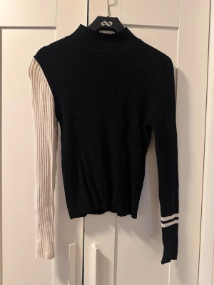 Svart stickad tröja med kontrastärm - Trendig ribbstickad tröja i svart med en beige kontrastärm och två vita ränder på ena ärmen. Hög hals och långärmad modell i mjukt, stretchigt material. Storleken är L men är liten i passformen, storlek M!