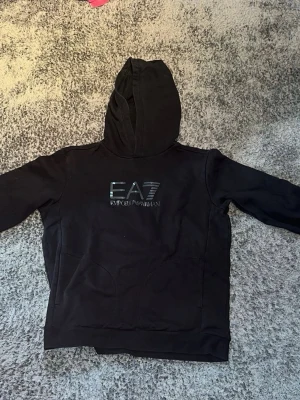 Svart hoodie från EA7 Emporio Armani S Herr - Säljer en svart hoodie från EA7 Emporio Armani med logga på bröstet. Hoodien har huva med dragsko och en stor magficka framtill. Tillverkad i mjukt material och har en clean, stilren look som passar perfekt till streetwear.