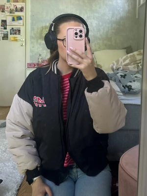Svart bomberjacka med beige ärmar - Säljer en oversized svart bomberjacka med beige ärmar och broderad text 'MG' på bröstet samt '1990' i rött på ryggen. Jackan har ribbade muddar och krage med vita ränder. Perfekt för dig som gillar retrostil och streetwear.