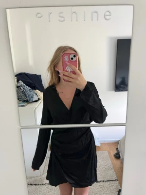 Svart omlottklänning från H&M - Snygg svart omlottklänning från H&M med v-ringning och långa ärmar. Klänningen har en draperad detalj vid midjan och är tillverkad i ett mjukt, lätt material som ger en elegant look. Perfekt för dig som gillar stilrena och trendiga plagg.