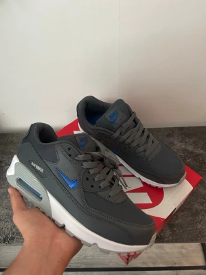 Nike Air Max 90 grå/blå sneakers - Snygga Nike Air Max 90 sneakers i mörkgrått med blå detaljer och vit sula. Skorna har en klassisk låg siluett, mesh och syntetmaterial i ovandelen samt den ikoniska Air Max-dämpningen i hälen. Perfekta för dig som gillar streetwear och sportig stil.