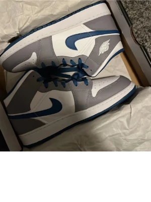 Nike Air Jordan 1 Mid grå/blå - Snygga Nike Air Jordan 1 Mid sneakers i grått och vitt med blå detaljer och blå sula. Klassisk design med ikonisk vinge-logga på sidan och blå swoosh. Ovandel i läder och textil, perfekt för dig som gillar streetwear och sportig stil.