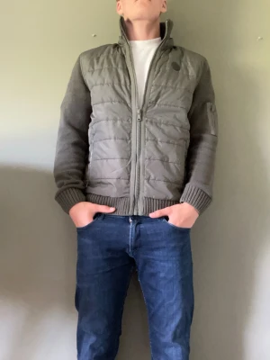 Cardigan - Snygg grön cardigan med stickade ärmar. Jackan har hög krage, dragkedja framtill och ribbade muddar. Perfekt för dig som vill ha en stilren och bekväm look. Finns också luva.