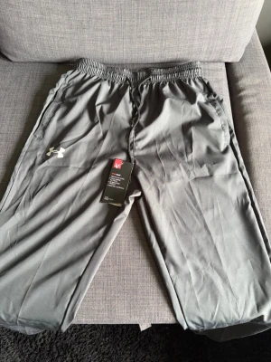 Grå polyester byxor från Under Armour - Snygga grå polyester byxor från Under Armour med dragsko i midjanoch sidofickor med dragkedja . Byxorna har en avslappnad passform och är tillverkade i ett mjukt, lätt material som känns skönt mot huden. Perfekta för chill eller träning.