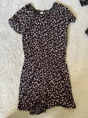 Blommig svart jumpsuit med kort ärm - Söt svart jumpsuit med små lila och rosa blommor, korta ärmar och resår i midjan för en skön passform. Tunt, mjukt tyg som känns luftigt och perfekt för varma dagar. Blommigt mönster ger en trendig och ungdomlig vibe.