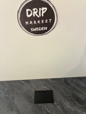 Svart korthållare från Sandlund hossein - Minimalistisk svart korthållare från Sandlund hossein i slätt läder. Klassisk design med plats för flera kort och diskret logotyp präglad på framsidan. Perfekt för dig som vill ha en stilren och smidig accessoar i fickan.