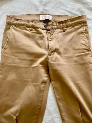 Beige chinos från These Glory Days - Snygga beige chinos från These Glory Days med klassisk rak passform. Byxorna har framfickor, två bakfickor med knapp och bälteshällor. Tillverkade i mjukt bomullstyg som ger en clean och stilren look. Mycket bra skick, framstår nästintill som nya. 