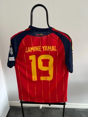 Spansk landslagströja Lamine Yamal - Säljer en officiell röd Spanien fotbollströja från Adidas med gula detaljer och nummer 19, Lamine Yamal på ryggen. Tröjan har blå ärmar, tunna gula ränder och broderad landslagssköld på bröstet. Perfekt för match eller träning. Vi har de flesta vm tröjorna hemma och borta. Mycket hög kvalitet och autentiska vuxen tröja  och barn set. Till det facila priset av 299kr. 