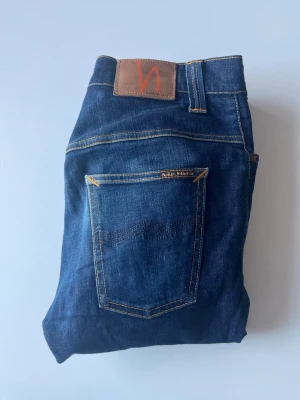 Mörkblå jeans från Nudie Jeans - Säljer ett par blå jeans från Nudie Jeans med klassisk femficksdesign och brunt läderpatch bak. Jeansen har en slim fit och är tillverkade i väldigt rå denim. Storleken är 30/32 men sitter lite kortare än så. Skriv såklart vid frågor eller funderingar🙌