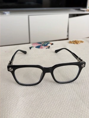 Chrome hearts glasögon - Snygga svarta glasögon med rektangulär form och chrome hearts silverdetaljer. Bågarna är i plast och har en stilren look som passar till det mesta. Perfekta för dig som vill ha en klassisk men ändå trendig accessoar. Det är ingen styrka i glasögonen så vem som helst kan använda dem