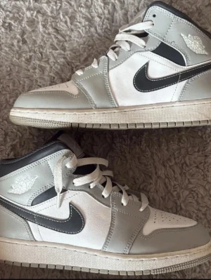 Nike Air Jordan 1 Mid grå/vit - Snygga Nike Air Jordan 1 Mid sneakers i grått och vitt med svarta detaljer och klassisk swoosh. Skorna har perforerad tå, vita skosnören och ikoniska Air Jordan-loggan på sidan. Ovandelen är i läder och textil för en clean och sportig look.
