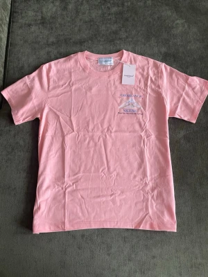 Rosa t-shirt från Casablanca - Casablanca t-shirt i ljusrosa bomull med tryck på bröstet och stort motiv på ryggen med texten 'Skiing' och berg. Klassisk passform och rund hals. Perfekt för dig som gillar coola prints och sportiga vibes.