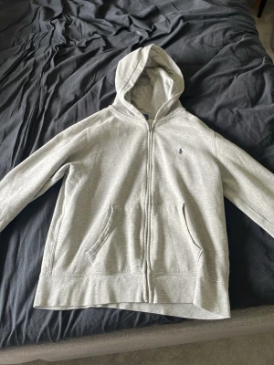 Grå zip hoodie från Polo Ralph Lauren - Snygg grå hoodie med dragkedja från Polo Ralph Lauren. Klassisk design med huva, två fickor framtill och den ikoniska blå broderade loggan på bröstet. Tillverkad i mjukt bomullsmaterial som är skönt att ha på sig. Perfekt för en stilren look.