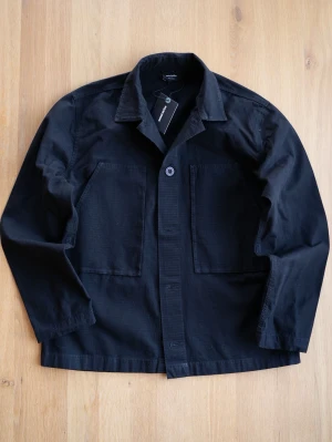 Ny ripstop Overshirt, size M - Stilren ny svart overshirt från Sweet med klassisk krage och två stora bröstfickor. Tillverkad i ett slitstarkt bomullstyg med diskret rutstruktur. Perfekt lager-på-lager-plagg med enkel knappstängning framtill.