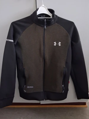 Svart och mörkgrå grön vindjacka Under Armour - Sportig vindjacka från Under Armour i svart och mörkgrått med dragkedja framtill och två fickor med dragkedja. Jackan har hög krage, lång ärm och logga på bröstet. Materialet är syntet och polyester, perfekt för aktiva dagar.