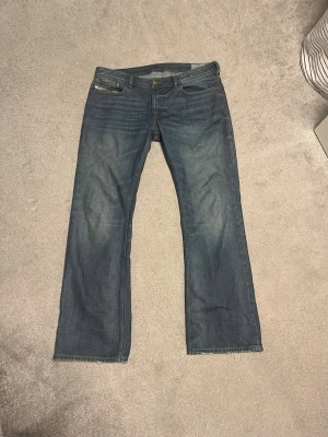 Blå bootcut jeans med slitningar - Säljer ett par klassiska blå jeans med bootcut passform och lätt slitna detaljer. Jeansen har fem fickor, knappgylf och är tillverkade i denim. Perfekta för en avslappnad och stilren look.