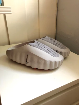 Grå Nike tofflor med vågig sula - Säljer ett par grå tofflor från Nike med unik vågig sula och ventilerande hål på ovansidan. Tofflorna har en modern design med diskret Nike-logga på sidan och orange detaljer på insidan. Tillverkade i ett lätt och slitstarkt syntetmaterial.