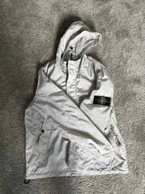 Stone Island ljusgrå vindjacka - Ljusgrå vindjacka från Stone Island med klassisk logopatch på vänster ärm. Jackan har huva, dragkedja och knappstängning samt två fickor med knappar framtill. Materialet är lätt och krispigt, perfekt för att skydda mot vind. Snygg och stilren design. Pris kan alltid diskuteras