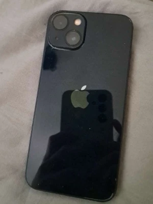 Svart iPhone 13 85% batterihälsa  - Snygg svart iPhone med stilren design och två kameror på baksidan. Telefonen har en glansig baksida med Apple-loggan och platt form. Perfekt för dig som gillar modern teknik och vill ha en mobil med bra kamera och klassisk Apple-känsla. telefonen fungerar utan problem och har hel skärm och baksida bara lite repor på sidan