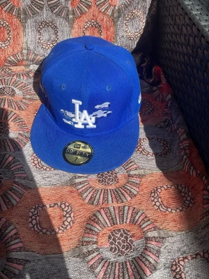 Blå LA Dodgers keps från New Era - Säljer en blå fitted keps från New Era, modell 59FIFTY, med LA Dodgers-logga framtill och MLB-logga bak. Kepsen har platt skärm och är gjord i ett tåligt tygmaterial. Perfekt för dig som gillar streetwear och sportig stil.