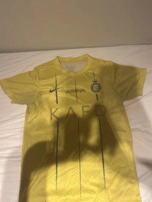 Gul Al Nassr matchtröja Nike - Snygg gul matchtröja från Al Nassr FC med blå detaljer och klubbmärke på bröstet. Tröjan har rund hals, korta ärmar och tryckta loggor från Nike och KAFD. Tillverkad i lätt och ventilerande material, perfekt för fotboll eller träning.