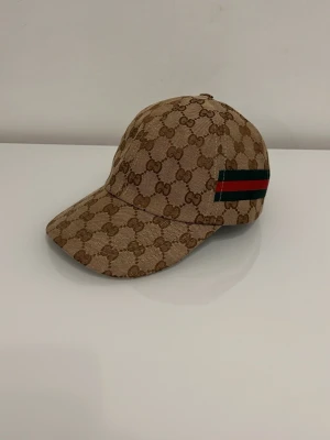 Beige keps - Säljer en beige keps  med det klassiska monogrammet över hela kepsen. På sidan finns en grön och röd rand som ger extra lyxig känsla. Kepsen har böjd skärm .