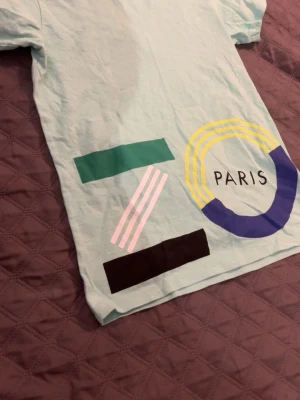 Kenzo Paris grafisk t-shirt - Snygg ljusblå t-shirt från Kenzo med färgglada grafiska tryck och texten 'PARIS' på framsidan. T-shirten har korta ärmar och är tillverkad i mjuk bomull. Perfekt för dig som gillar statement-plagg och streetstyle.