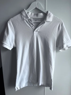 Vit pikétröja från Dressmann - Klassisk vit pikétröja från Dressmann med krage och knappar framtill. Tröjan har korta ärmar och är tillverkad i bomull, perfekt för en clean och stilren look.