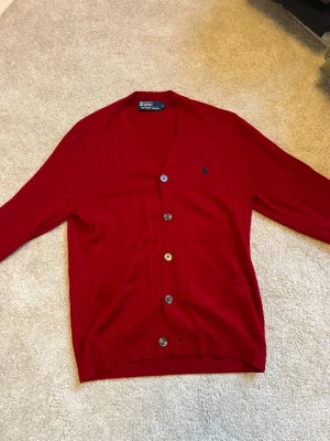 Röd cardigan från Polo Ralph Lauren - Snygg röd cardigan från Polo Ralph Lauren med knappar framtill och två fickor. Klassisk blå logga broderad på bröstet. Tillverkad i mjukt material och har ribbade muddar vid ärmslut och nederkant. Helt ny och aldrig använd. Till och med reserv knapparna sitter fast fortfarande 