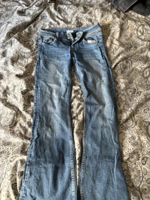 Blå bootcut jeans med fickdetaljer - Säljer ett par ljusblå bootcut jeans med snygga fickdetaljer både fram och bak. Jeansen har klassisk femficksdesign, knappgylf och kontrastsömmar. Perfekta för dig som gillar en avslappnad men trendig look.