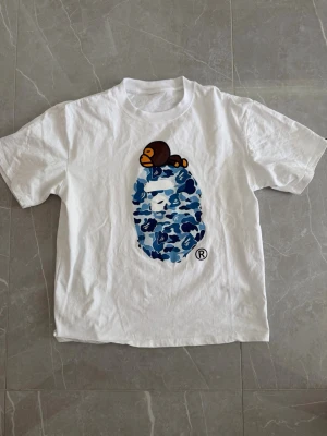 Vit BAPE t-shirt med blått camomönster - Säljer en vit t-shirt från BAPE med ett stort blått camomönstrat tryck på bröstet och en liten apa ovanpå. T-shirten har rund hals och korta ärmar. Perfekt för dig som gillar streetwear och vill sticka ut med en unik design.