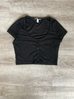 Svart croppad topp från H&M - Snygg svart croppad topp från H&M med korta ärmar och knappar framtill. Ganska form sittande. Toppen har ett diskret hålmönster i tyget och en djup rundad urringning. 