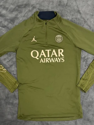 PSG Jordan träningströja - Säljer en olivgrön långärmad träningströja från PSG och Jordan med Qatar Airways-tryck på bröstet. Tröjan har halv dragkedja, diskret mönster på ärmarna och Paris Saint-Germain-logga. Perfekt för fotbollsträning eller chill. 