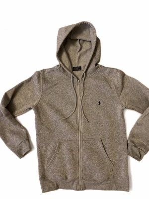 Grå zip hoodie från Polo Ralph Lauren - Klassisk grå hoodie med dragkedja från Polo Ralph Lauren. Tröjan har huva med dragsko, två fickor framtill och den ikoniska lilla broderade loggan på bröstet. Tillverkad i mjuk bomullsblandning för extra komfort.