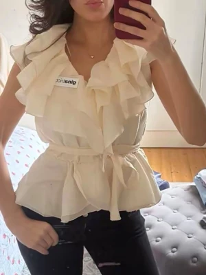 Krämvit volangblus från Gina Tricot - Säljer en krämvit blus från Gina Tricot med volanger och knytskärp i midjan. Aldrig använd endast testad! Blusen är ärmlös och har en lätt skimrande, luftig materialkänsla som ger ett elegant intryck. Perfekt för dig som gillar romantiska detaljer och en feminin look. 