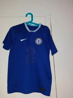 Chelsea FC blå matchtröja Nike - Säljer en blå Chelsea FC matchtröja från Nike med klubbens emblem på bröstet och sponsorlogga i mitten. Tröjan har korta ärmar och vit-blå detaljer runt kragen. Perfekt för fotbollsfans som vill visa sitt stöd på läktaren eller planen.