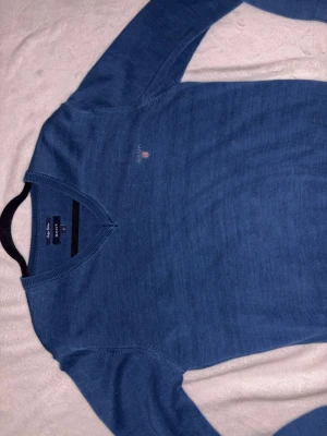 Blå V-ringad tröja från GANT - Snygg blå V-ringad tröja från GANT i Indigo Cotton. Tröjan har diskret logotyp broderad på bröstet och ribbade muddar vid hals, ärmslut och nederkant. Perfekt för dig som gillar klassisk och stilren design.