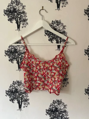 Blommig croppad linnetopp - Söt croppad linnetopp med blommigt mönster i rött, vitt och grönt. Toppen har tunna axelband och vågig kant längs halsen och nederkanten. Perfekt för dig som gillar färgglada och lekfulla plagg. Materialet känns lätt och somrigt.