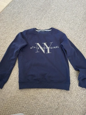 Mörkblå sweatshirt med tryck NY - Säljer en mörkblå sweatshirt med stort NY-tryck och texten 'ATHLETIC ORIGINAL' på bröstet. Tröjan har rund halsringning, långa ärmar och ribbade muddar vid ärmslut och nederkant. Perfekt för en avslappnad och sportig stil.
