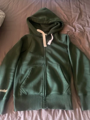 Mörkgrön hoodie med dragkedja - Mörkgrön hoodie med vit dragsko och dragkedja framtill. Modellen har en stor huva, ribbade muddar och två rymliga fickor på magen. Materialet är mjukt och fleeceliknande på insidan, perfekt för att hålla sig varm. Diskret broderad text på ärmen.