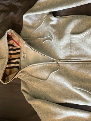 Grå hoodie från Burberry med dragkedja - Snygg grå hoodie från Burberry med klassiskt rutigt foder i huvan och silverfärgad dragkedja med Burberry-logga. Hoodien har två fickor framtill och är tillverkad i mjukt bomullsmaterial. Perfekt för dig som vill ha en stilren och exklusiv look.