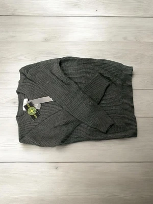 Grå stickad tröja från Stone Island - Snygg grå stickad tröja från Stone Island med klassisk rund hals och ribbade muddar. Tröjan har en diskret logga på armen och är tillverkad i ett mjukt, grovt stickat material som passar perfekt till höst och vinter.