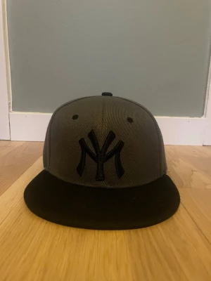 Grå New York Yankees keps - Snygg grå keps från New Era med svart NY Yankees-logga framtill och svart skärm. Klassisk snapback-modell med platt skärm och broderade ventilationshål. Perfekt accessoar för en sportig streetstyle-look.